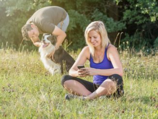 Mann streichelt Hund Frau sitzt im Gras und spielt mit dem Handy